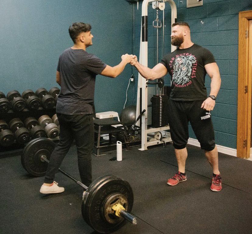 The photo depicts two men in a gym; one facing away from the camera, and the other facing towards it, smiling and engaging in a congratulatory handshake. The gym is equipped with various weights and exercise equipment, including a loaded barbell on the floor. Both men are dressed in workout attire, suggesting an active training session.